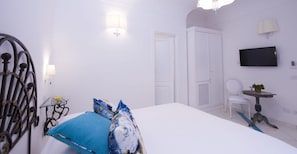 Une pièce blanche avec un lit, une chaise, une petite table avec une plante et un téléviseur fixé au mur.