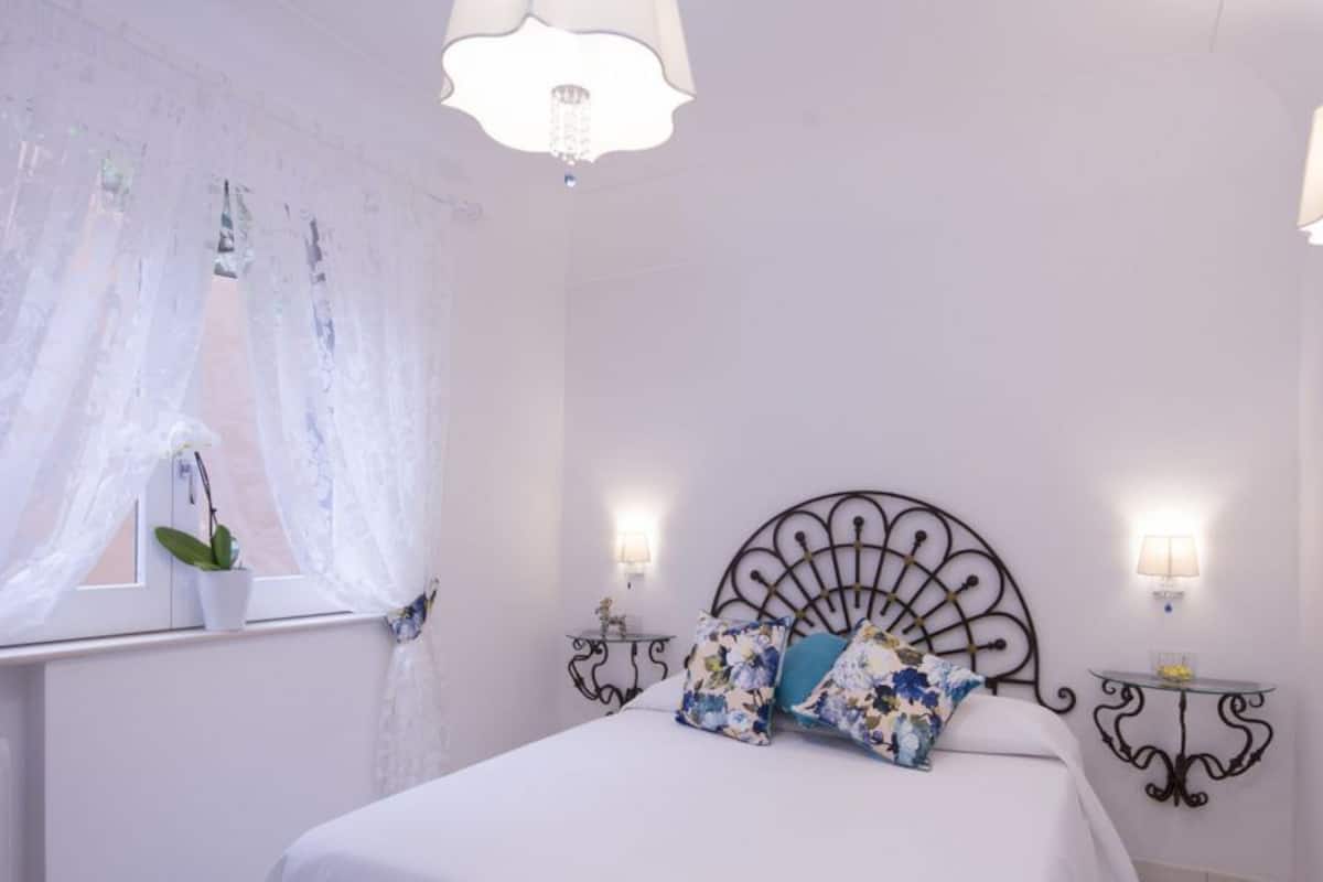 Un dormitorio con cama, ventana con cortinas translúcidas, planta en maceta y lámparas fijadas a la pared.