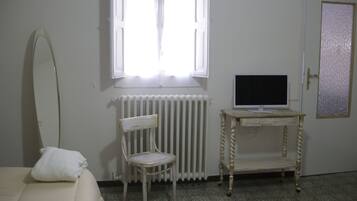 Ein Zimmer mit einem Bett, einem Sessel, einer Heizung, einem Fernseher auf einem kleinen Tisch und einer Tür mit einer Milchglasverglasung.