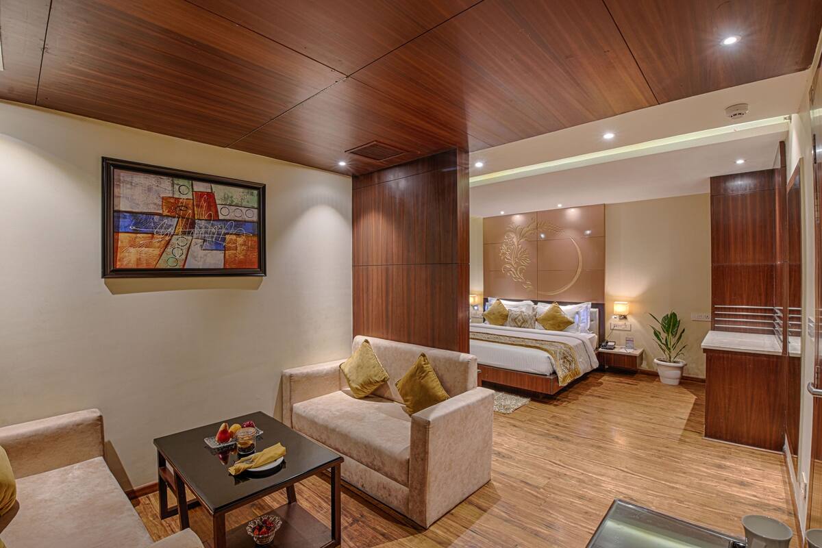 Habitación de hotel con techo de madera, una cama con mesita de noche, un sofá con cojines, una pequeña mesa con frutero y un cuadro en la pared.