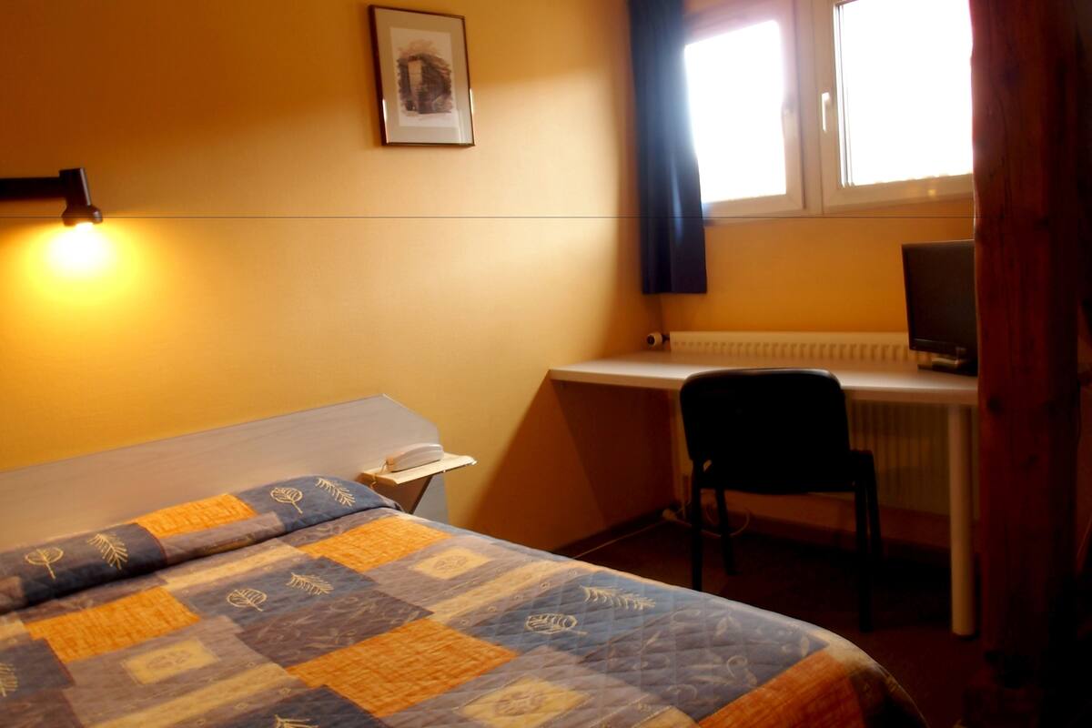A hotel room with a bed, a desk, a chair, a radiator, and a window with curtains.