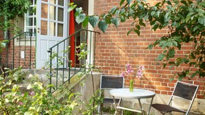 A brick building with a white door, a small outdoor seating area with a table and two chairs, and a potted plant with pink flowers. - Cy Twombly -. Villa-Hufeland in Berlin, quiet, 25 minutes by S-Bahn to the center (Berlin)