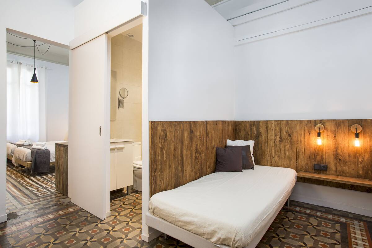 Un dormitorio con una cama, cabecera de madera, dos lámparas de pared, una mesita de noche y suelo estampado.