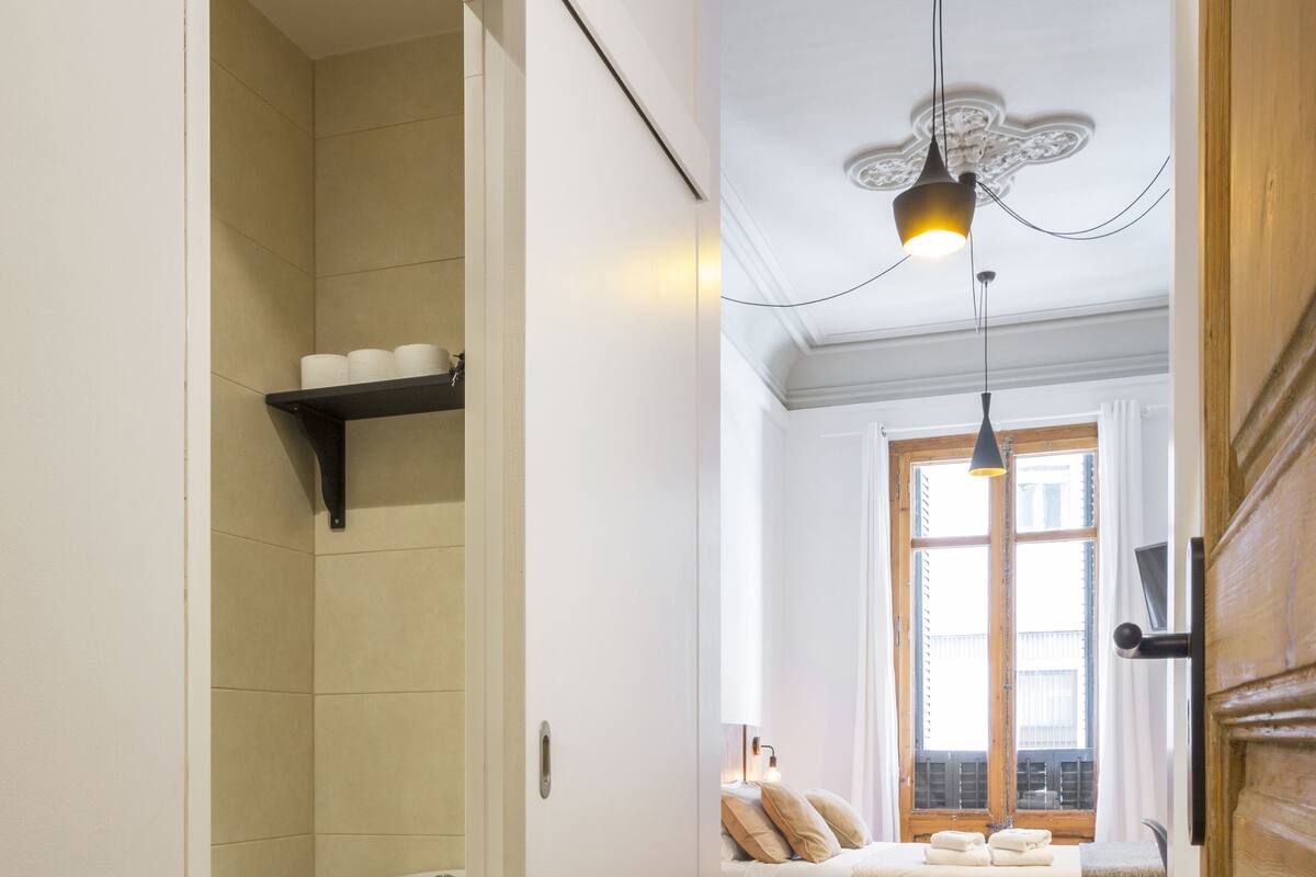 Un pasillo estrecho conduce a un baño con inodoro y ducha. En la habitación contigua hay una cama y un ventanal.