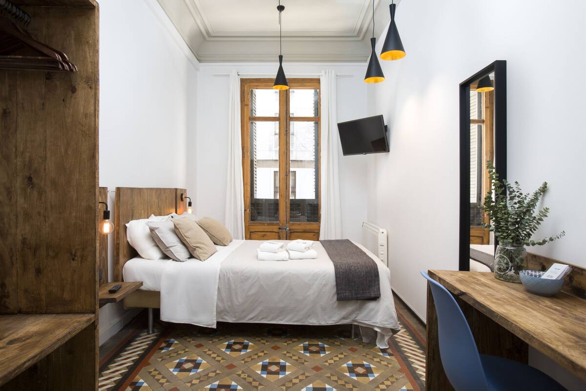 Un dormitorio con cama, cabecera de madera, mesitas de noche, un escritorio, una silla, televisor, espejo y ventana con vista a edificios.