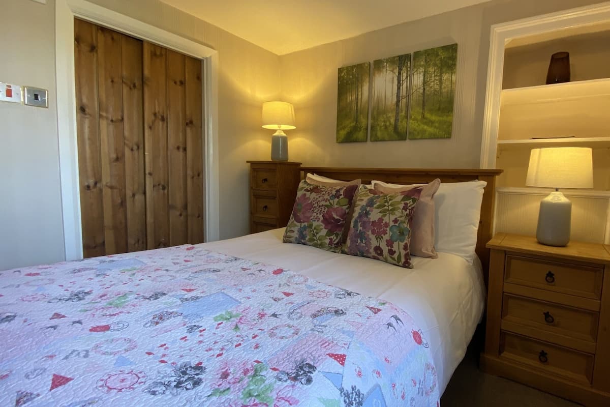 A bedroom with a bed, wooden wardrobe, two bedside tables with lamps, floral-patterned bedding, and a wall-mounted painting.