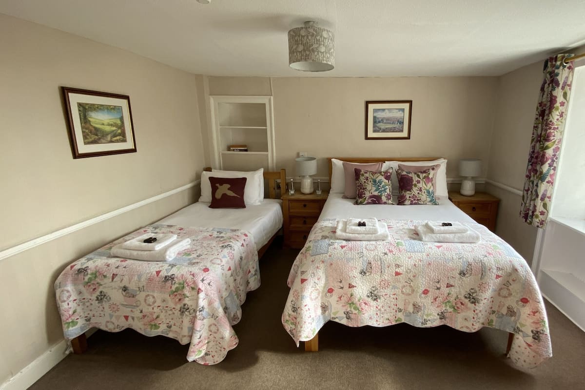 A room with two single beds, each with a floral bedspread and a pillow. There are bedside tables with lamps, a shelf above one bed, and a window with curtains.