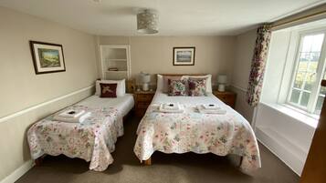 A room with two single beds, each with a floral bedspread and a pillow. There are bedside tables with lamps, a shelf above one bed, and a window with curtains.