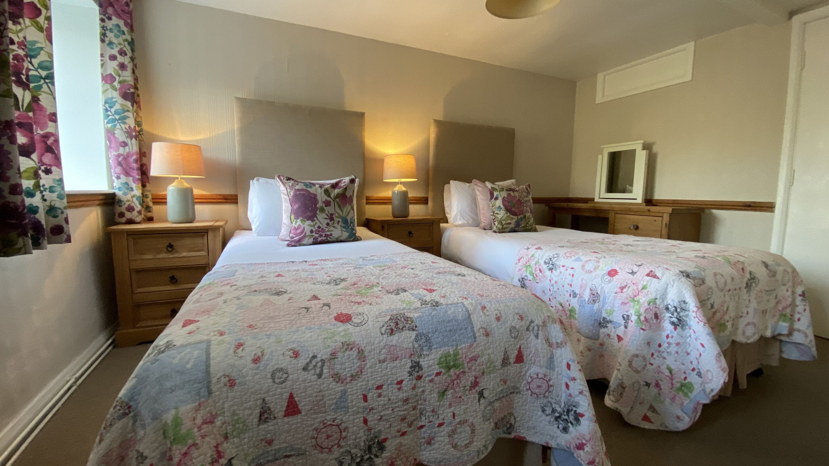 Habitación de hotel con dos camas, cada una con colcha estampada con motivos florales, lámparas de noche, mesitas de noche de madera y un espejo sobre el tocador.