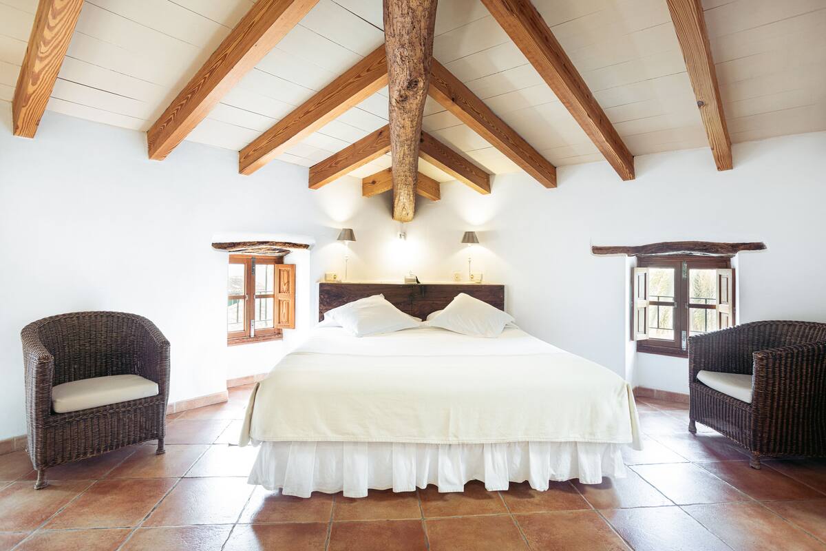 Quarto com uma cama grande, duas cadeiras de vime, vigas de madeira no teto e uma janela com persianas.