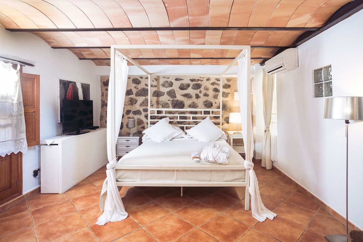 Quarto com parede de pedra, cama com roupa de cama branca, TV e escrivaninha com abajur.
