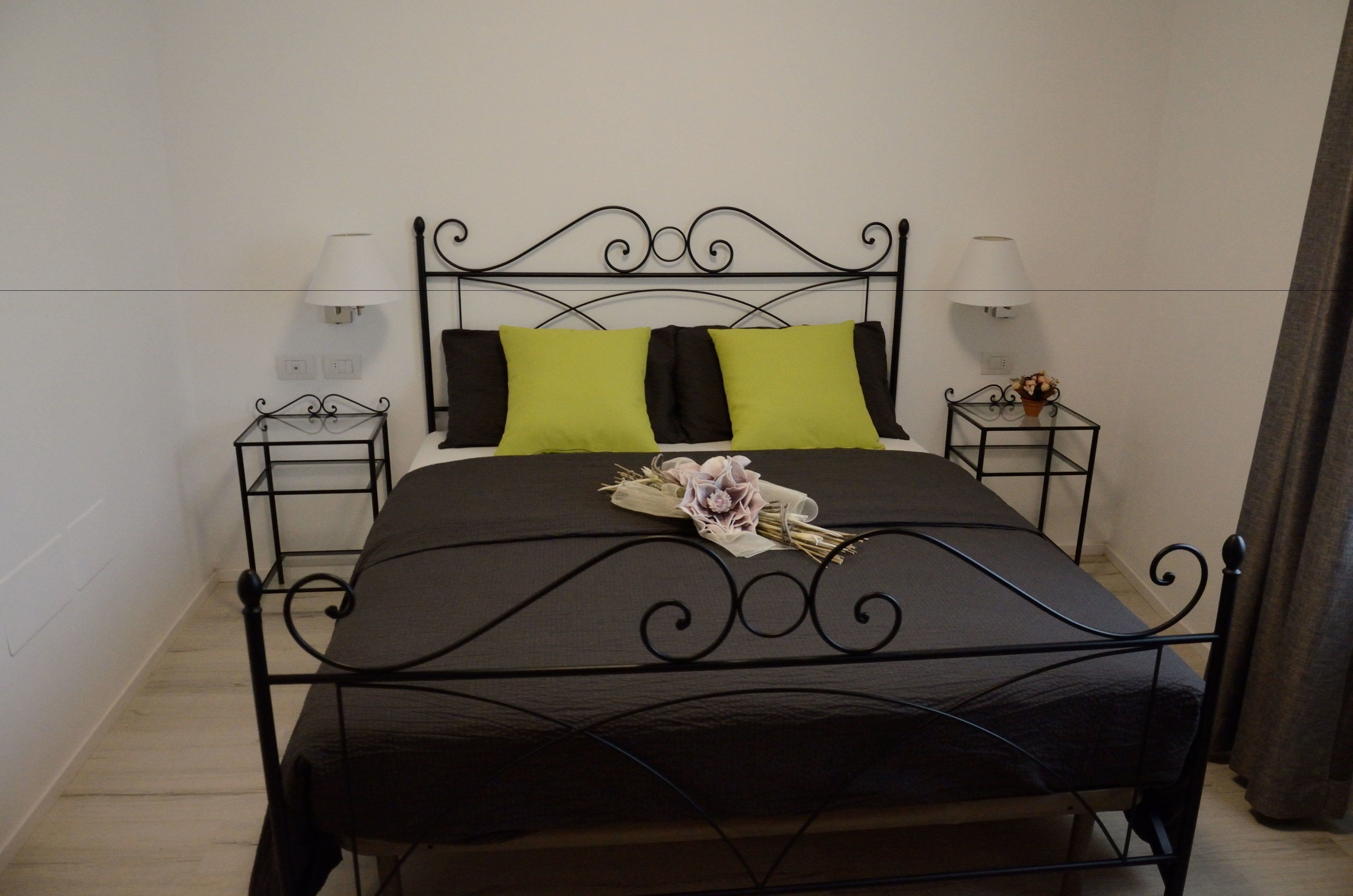 A bed with a black metal frame, two black pillows, and a green pillow. There are two bedside tables with lamps and a bouquet of flowers on the bed.