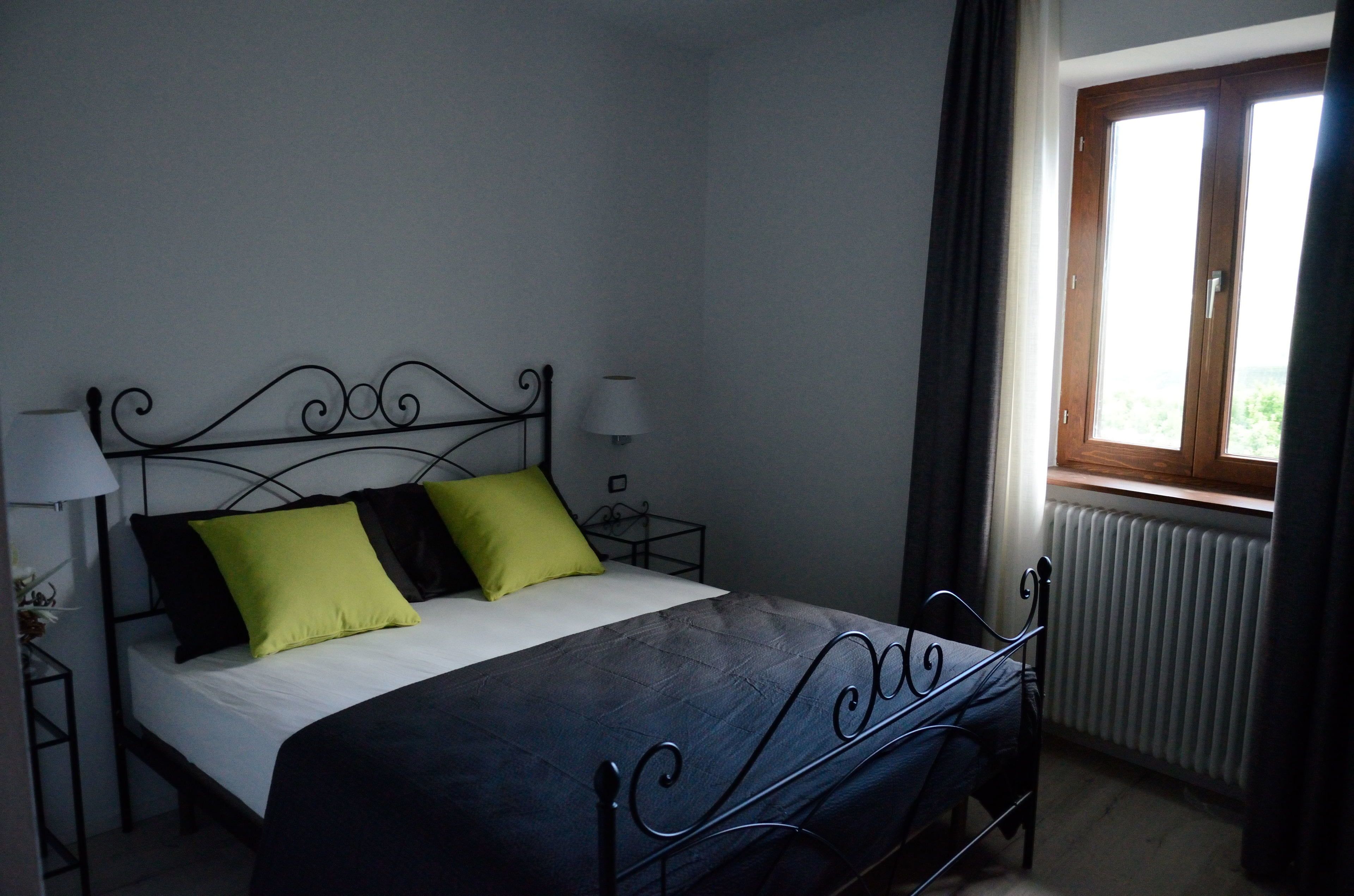 Un letto singolo con struttura in metallo, due comodini con lampade, una finestra con tende e un termosifone.