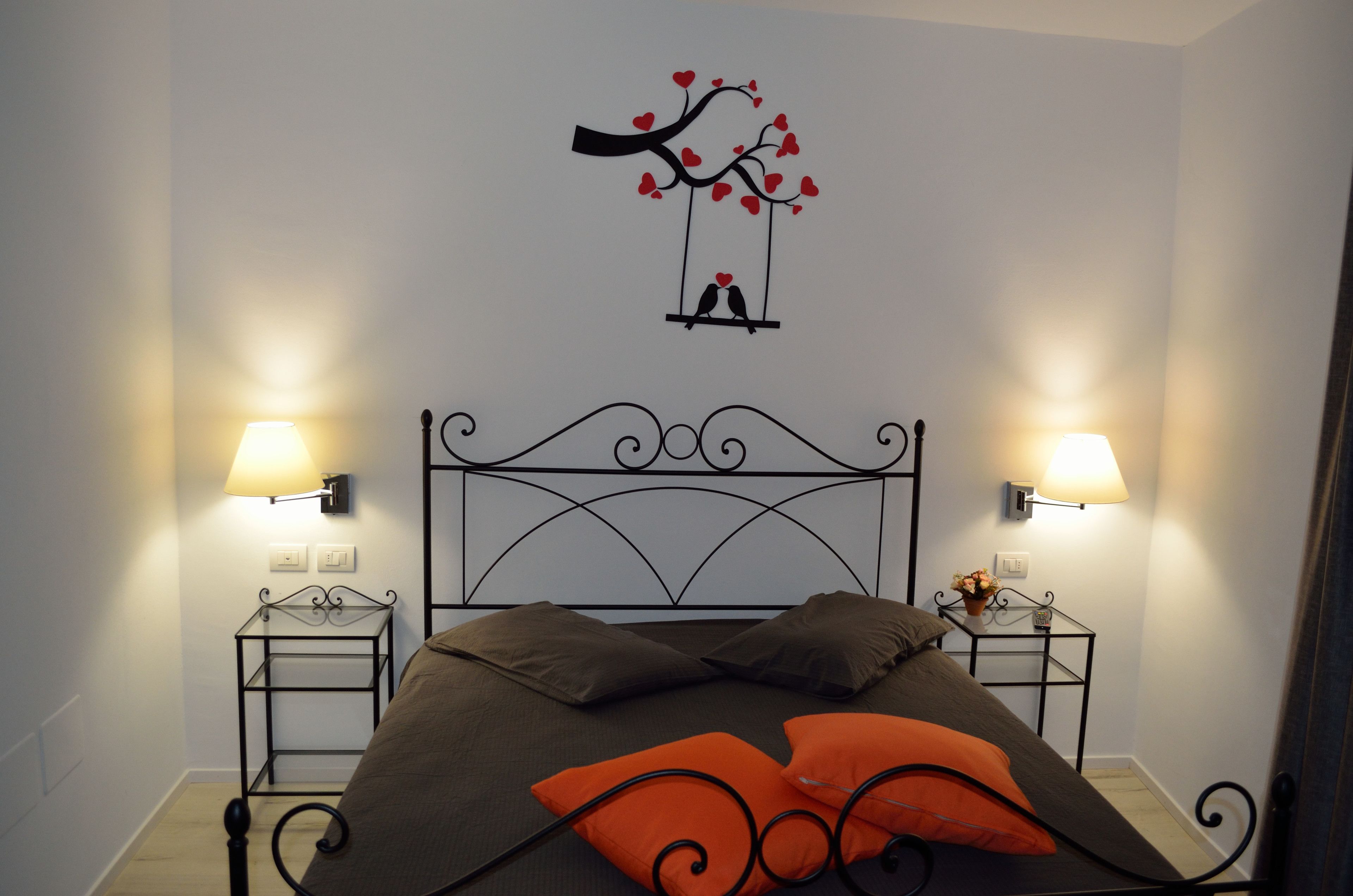 Una cama bien tendida con un cabecero, dos mesitas de noche con lámparas y un adorno de pared con un árbol y pájaros.
