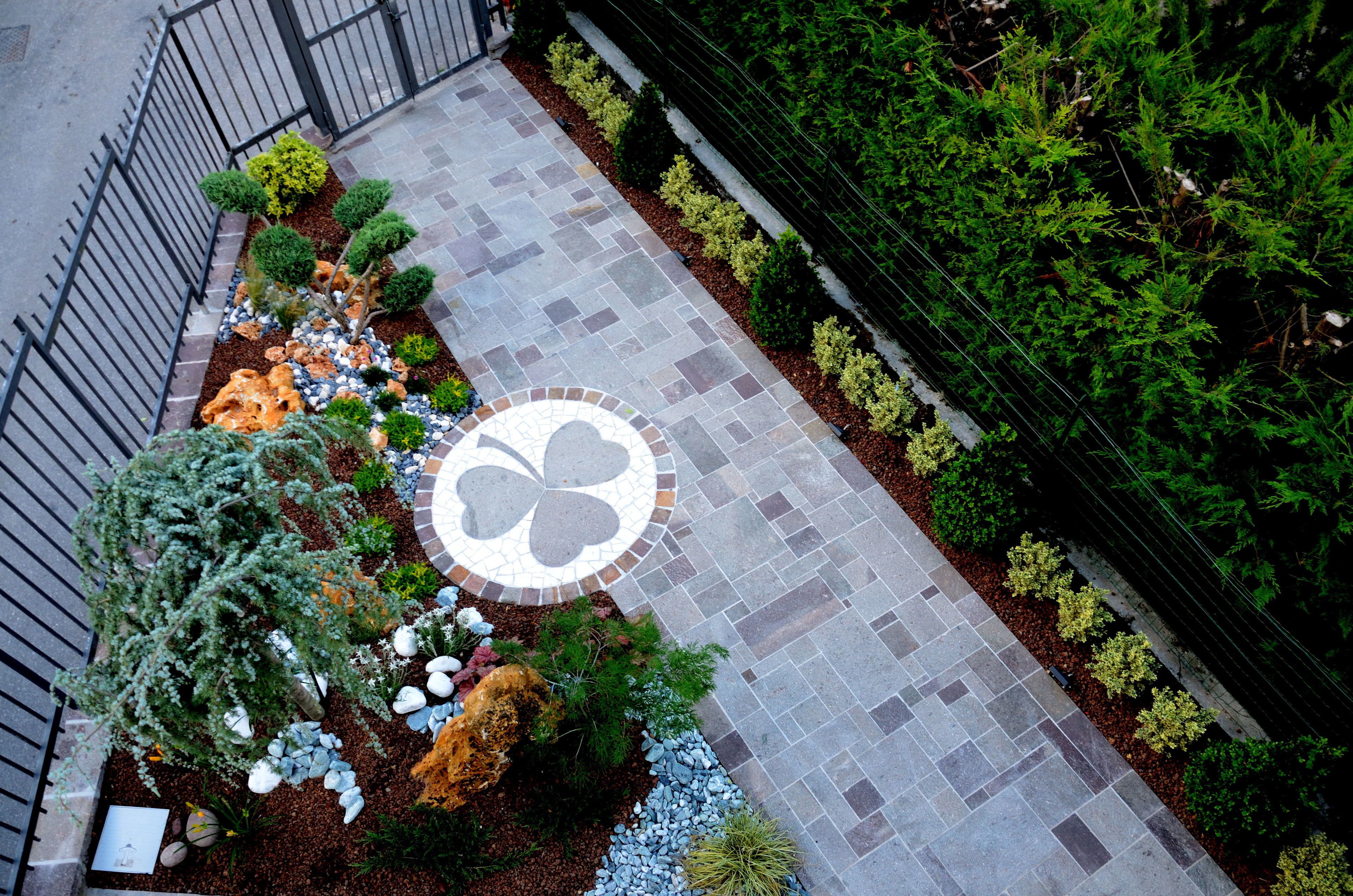 A landscaped pathway with a mosaic tile design, surrounded by various shrubs and small trees.