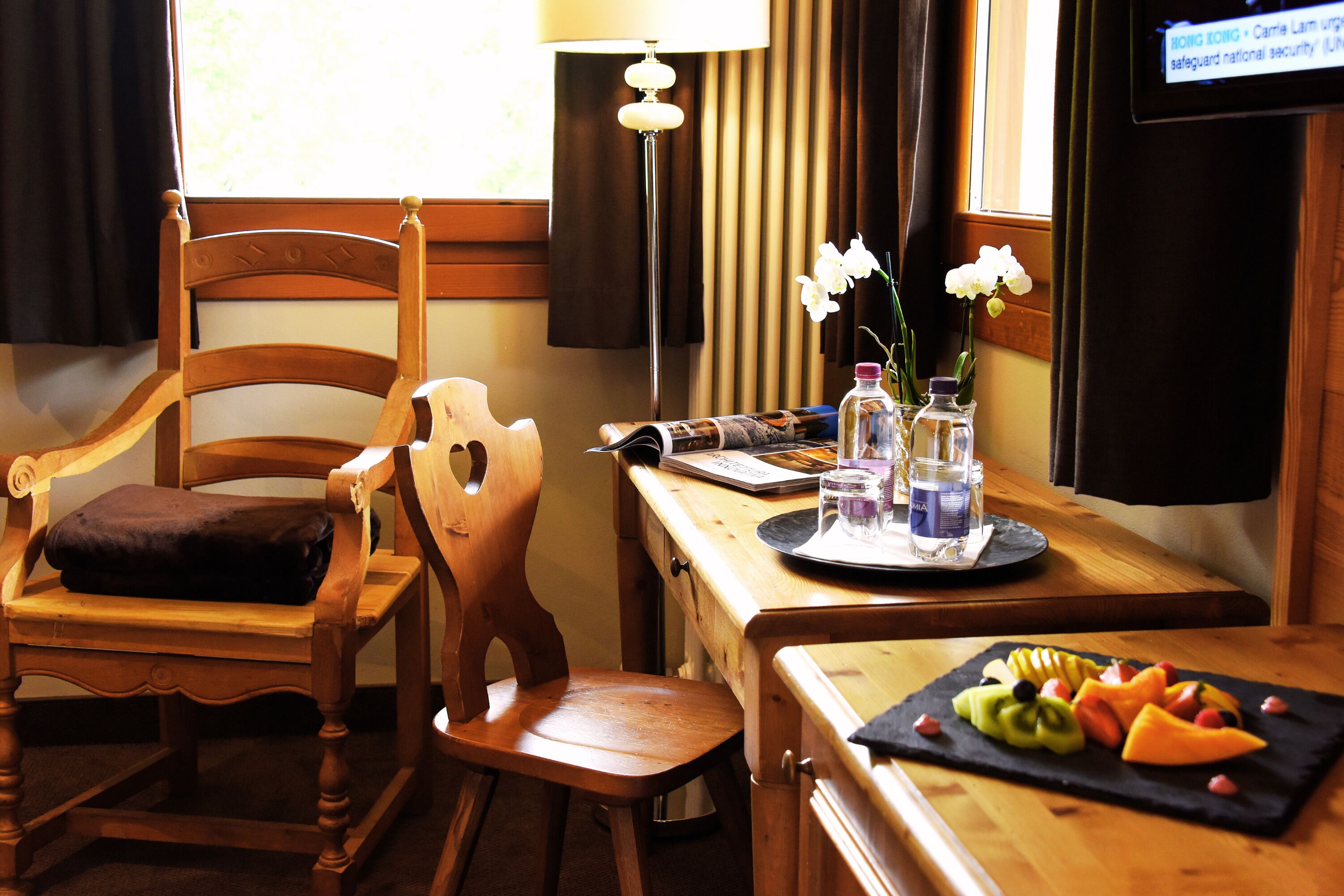 Une chaise en bois avec un coussin, une table en bois sur laquelle est disposée une assiette de fruits, une lampe et un écran de télévision affichant un message.