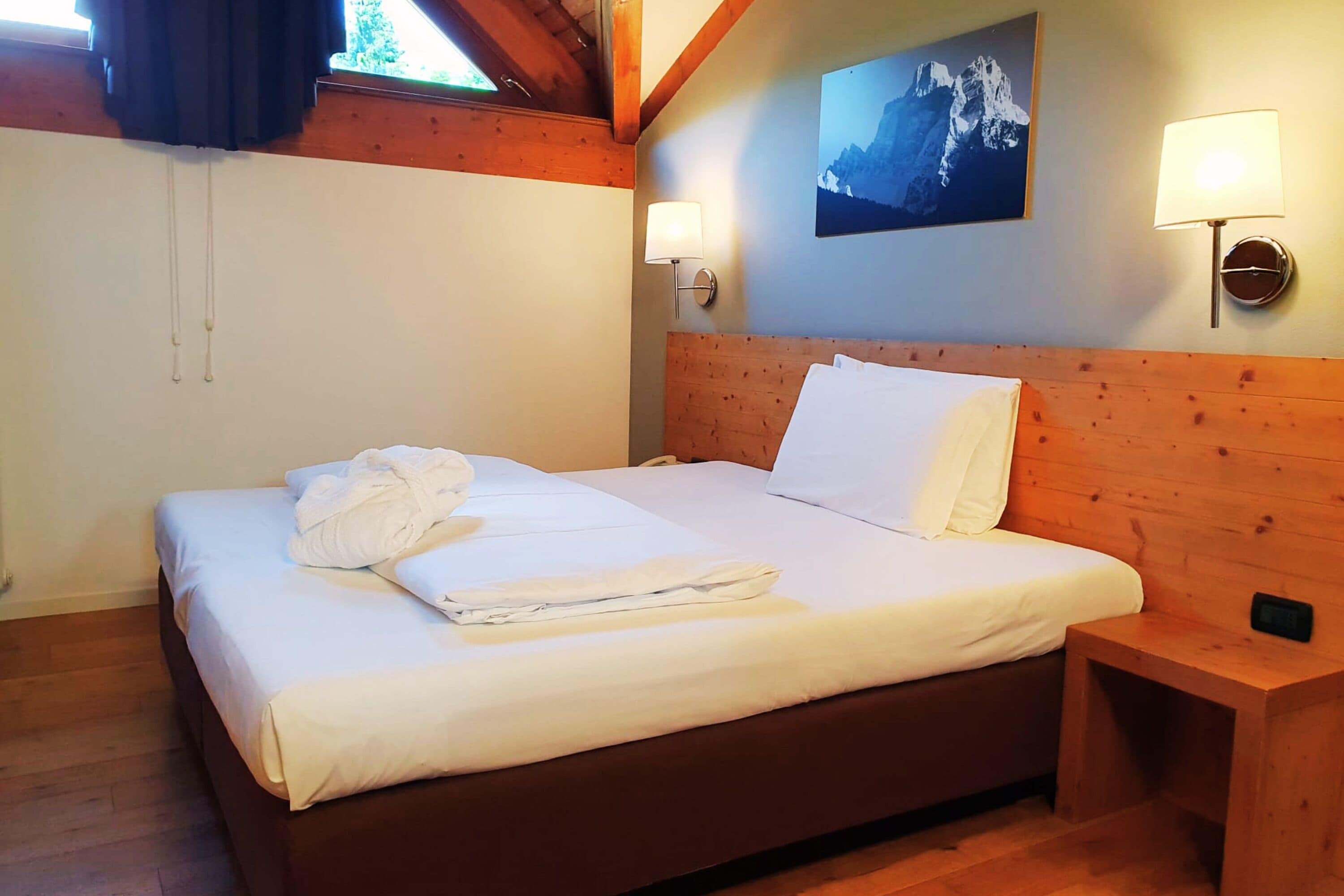 A neatly made bed with white linens and a folded towel, a wooden headboard, a nightstand with a lamp, and a mountain landscape painting on the wall.
