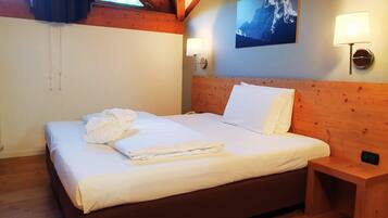 A neatly made bed with white linens and a folded towel, a wooden headboard, a nightstand with a lamp, and a mountain landscape painting on the wall.