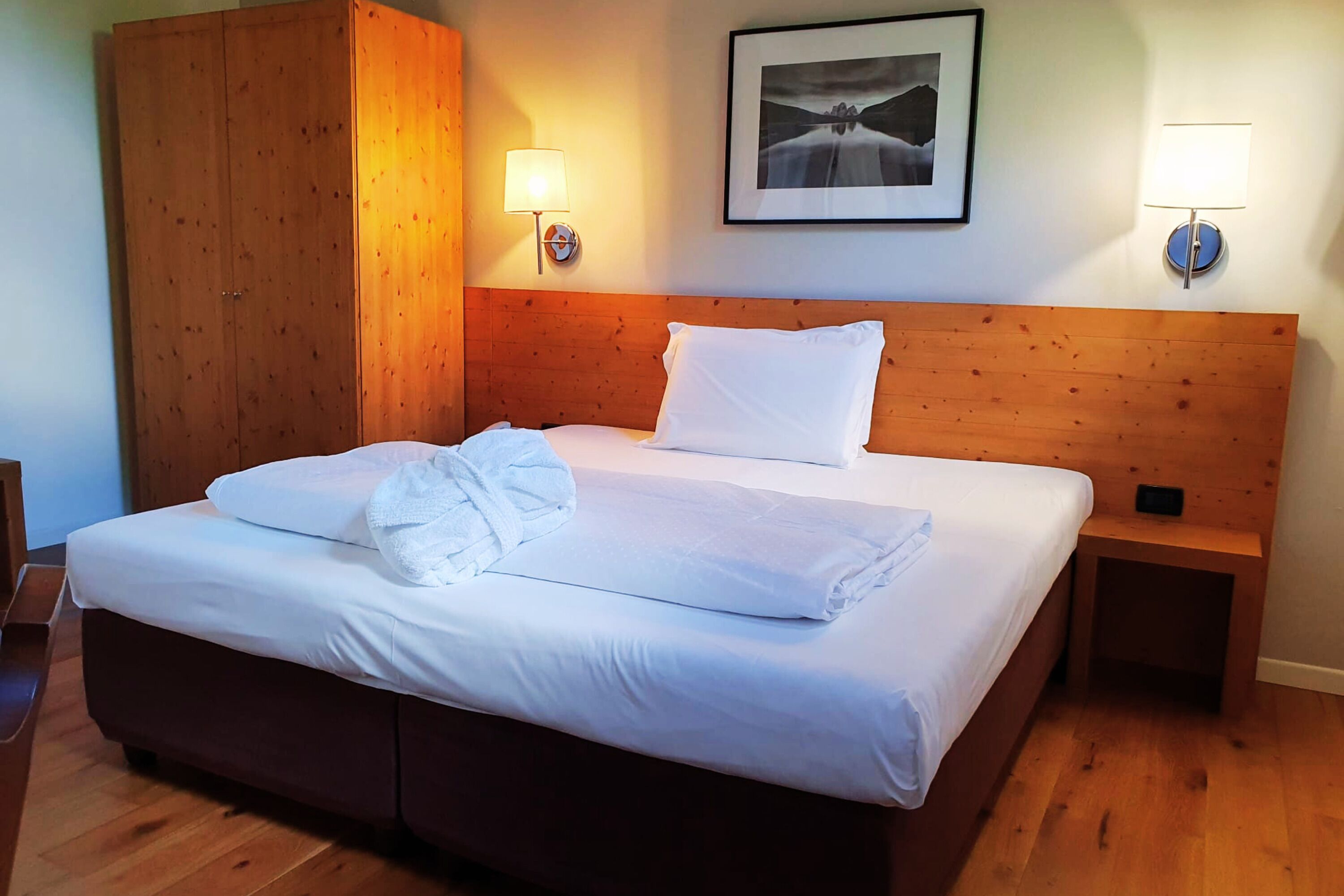 A neatly made bed with white linens and towels, a wooden headboard, two bedside lamps, and a framed picture on the wall.
