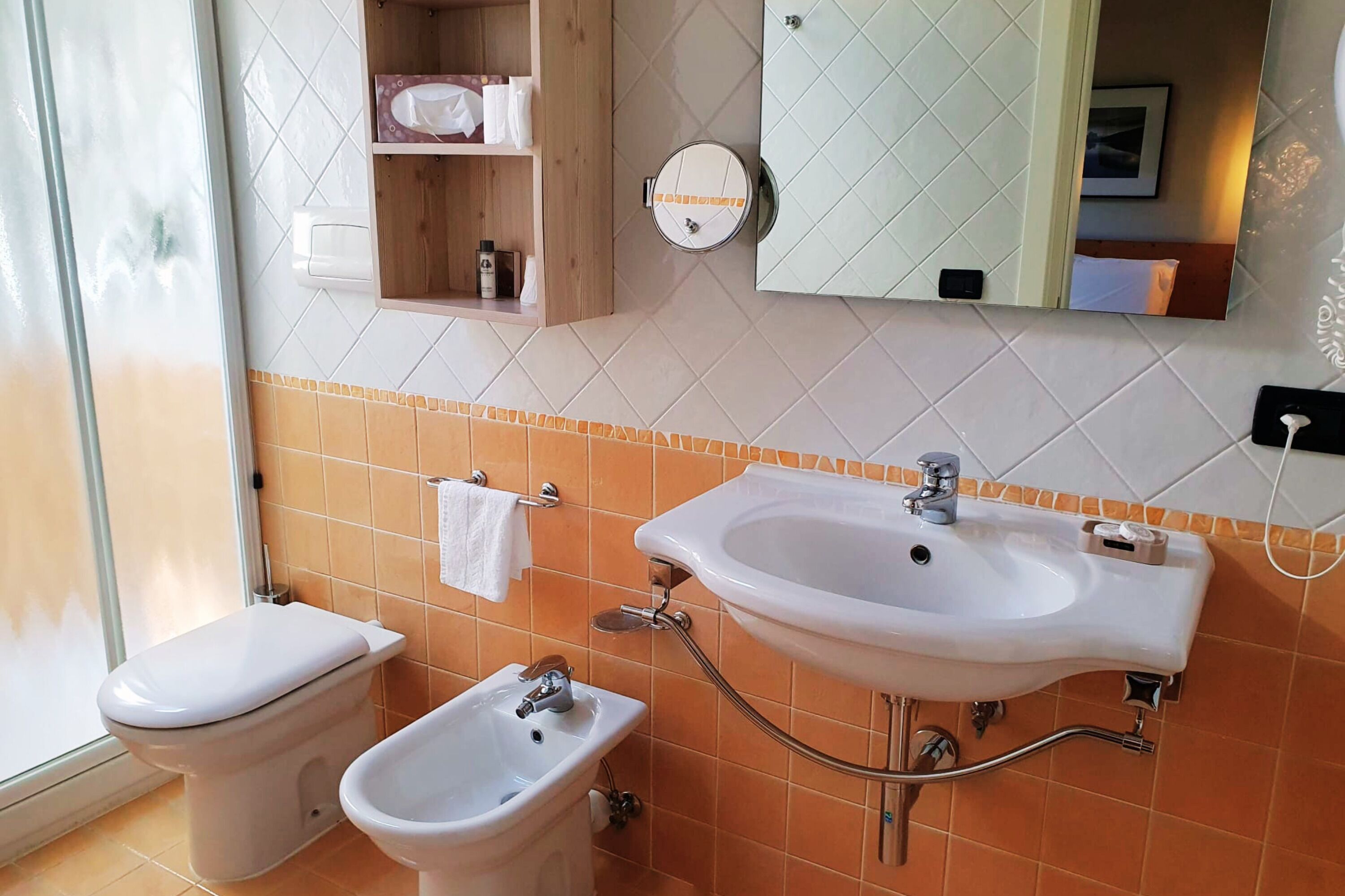 Casa de banho com sanita, bidé e lavatório, com um espelho sobre o lavatório e um prateleira sobre a sanita.