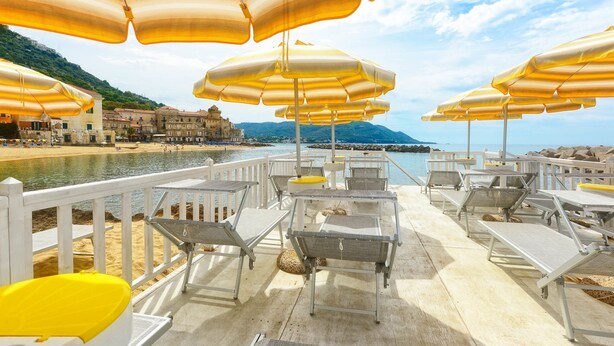 A seaside terrace with white lounge chairs and yellow umbrellas, overlooking a calm sea and a historic town.