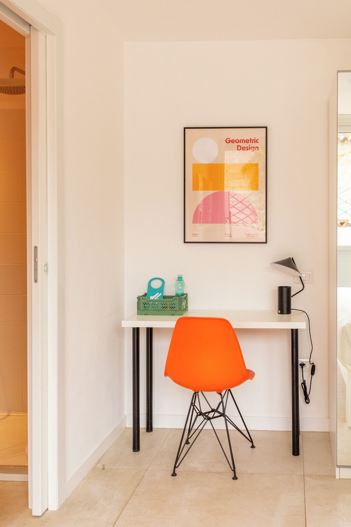 Un petit bureau comprenant un bureau, une chaise orange, une affiche encadrée présentant un motif géométrique et une caisse verte contenant divers objets.