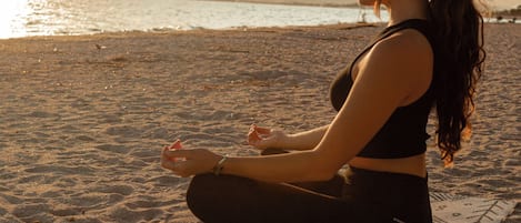 Una persona meditando sobre una esterilla de yoga en una playa, con vistas al mar y montañas al fondo.
