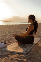Una persona meditando sobre una esterilla de yoga en una playa, con vistas al mar y montañas al fondo.