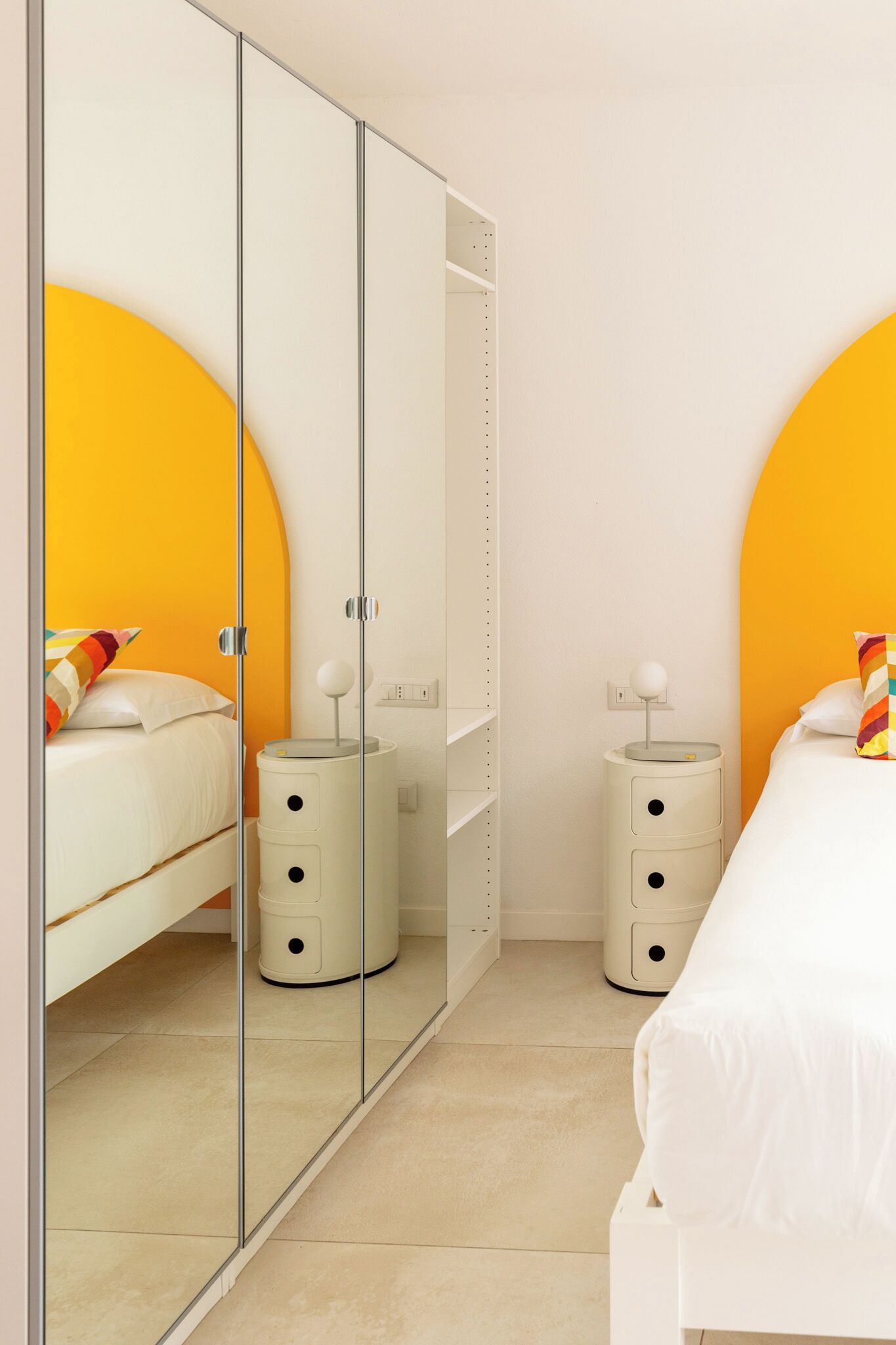 Un dormitorio con armario espejado, cama con ropa de cama blanca, dos mesitas de noche con lámparas y una pared de acento amarilla.