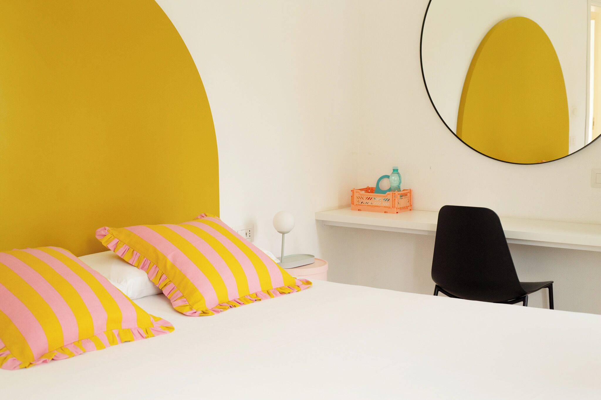 A bedroom with a bed, pillows, a desk, a chair, a mirror, and a wall-mounted shelf.