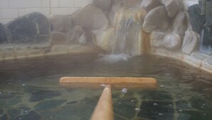 Una bañera de piedra con una paleta de madera, agua cristalina y un muro de azulejos con texto japonés.