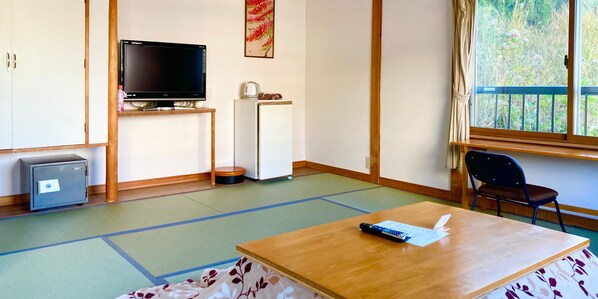 Una habitación con suelo de tatami, televisor, refrigerador, una caja de seguridad pequeña y ventana con vista a follaje.