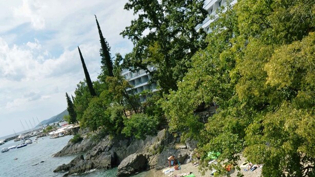 Ein Strand mit badenden und sonnenbächelnden Personen, umgeben von Bäumen und einer felsigen Küste.
