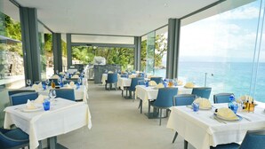 Ein Restaurant mit gedeckten Tischen, großen Fenstern mit Meerblick und einem Koch, der im Hintergrund kocht.