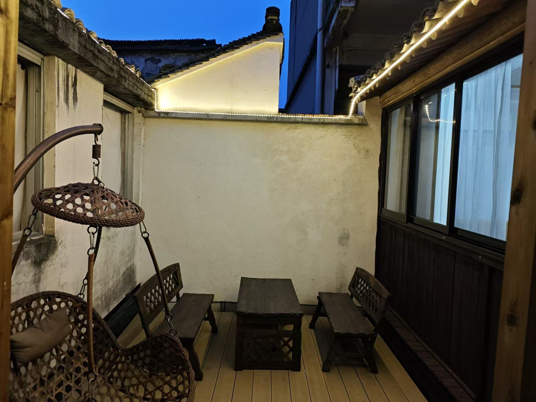 An outdoor patio with a wooden table and benches, a hanging chair, and a wall with a window.