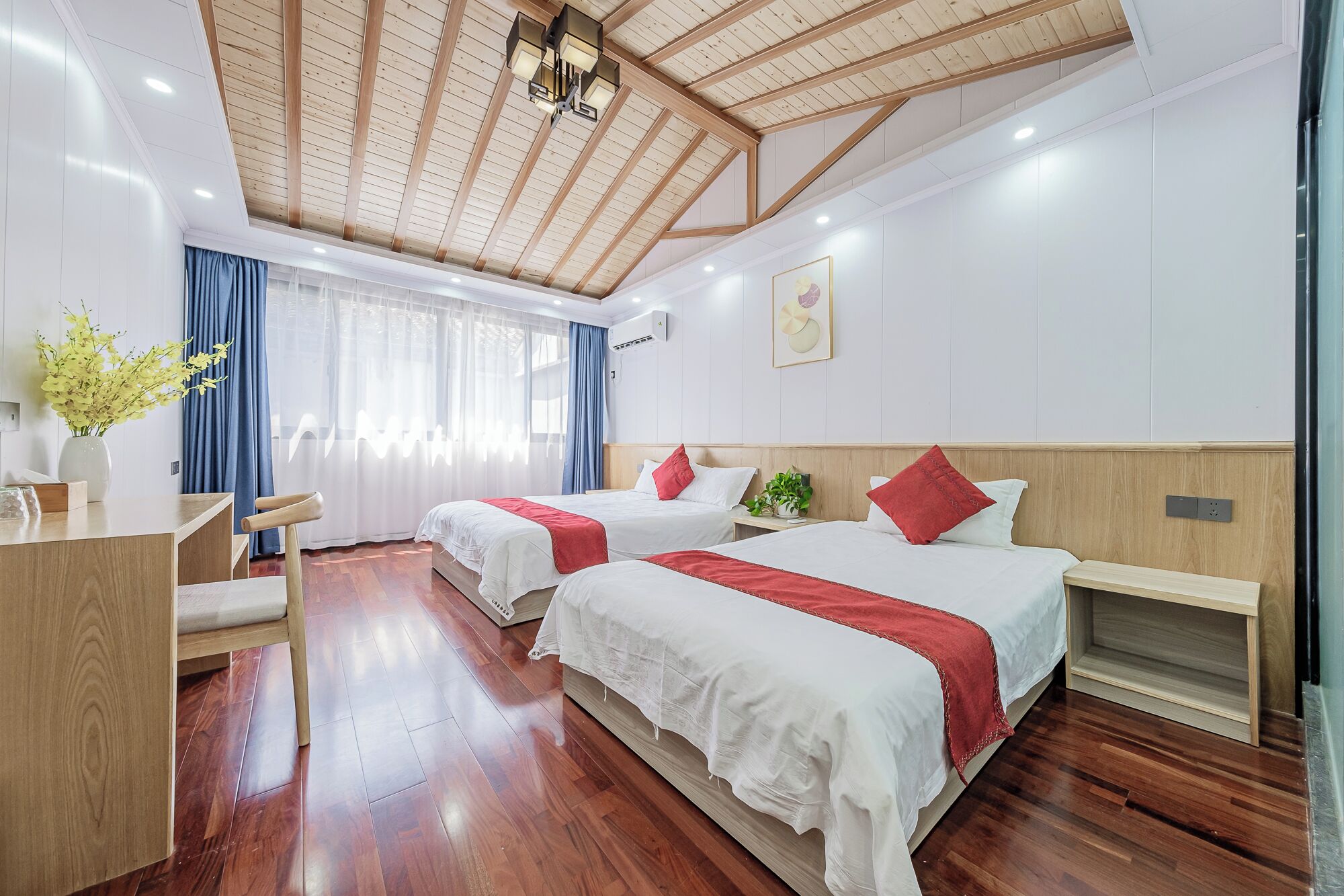 Camera d'albergo con due letti, pavimento in legno, una scrivania e un'ampia finestra con tende.