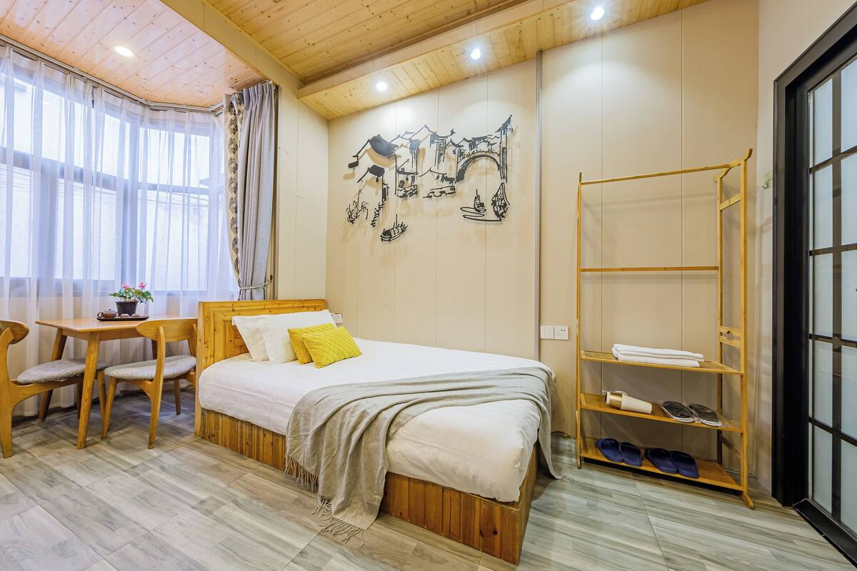 Un dormitorio con una cama de madera, una mesita con sillas, un estante en la pared y un ventanal con cortinas translúcidas.