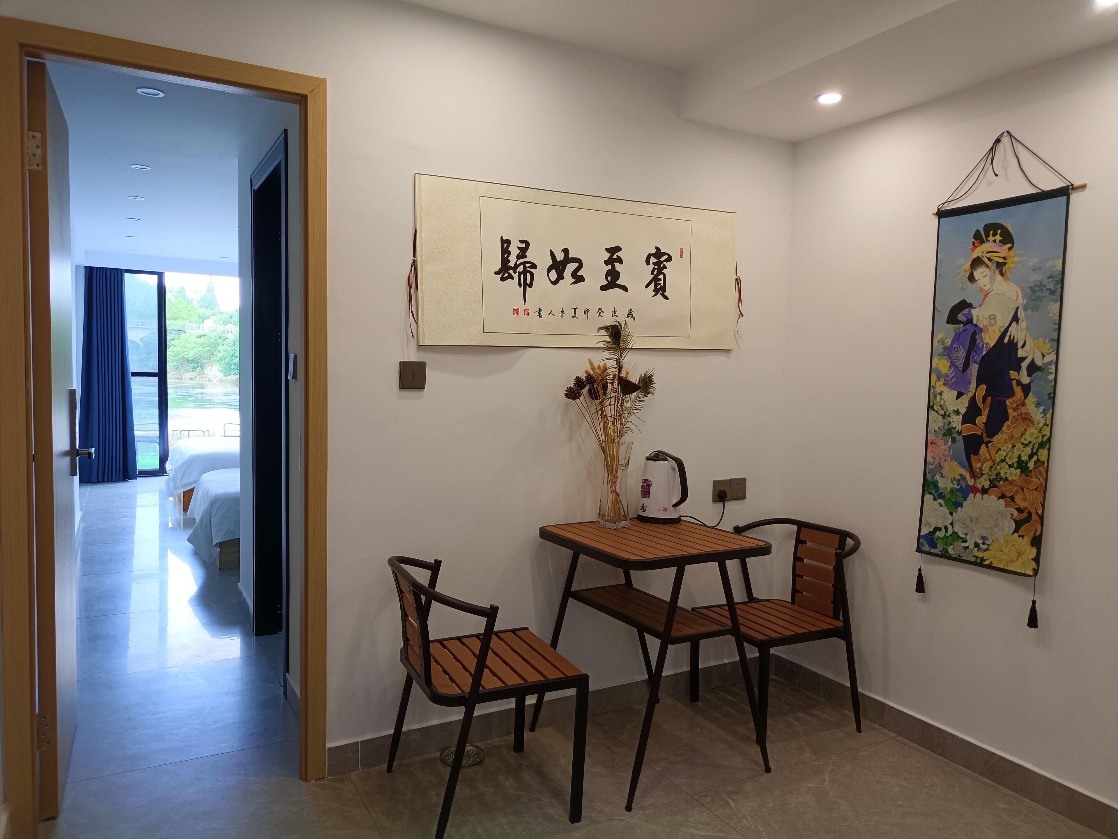 A hotel room with a bed, a table with chairs, a vase with dried flowers, a painting, and a sign with Chinese characters.
