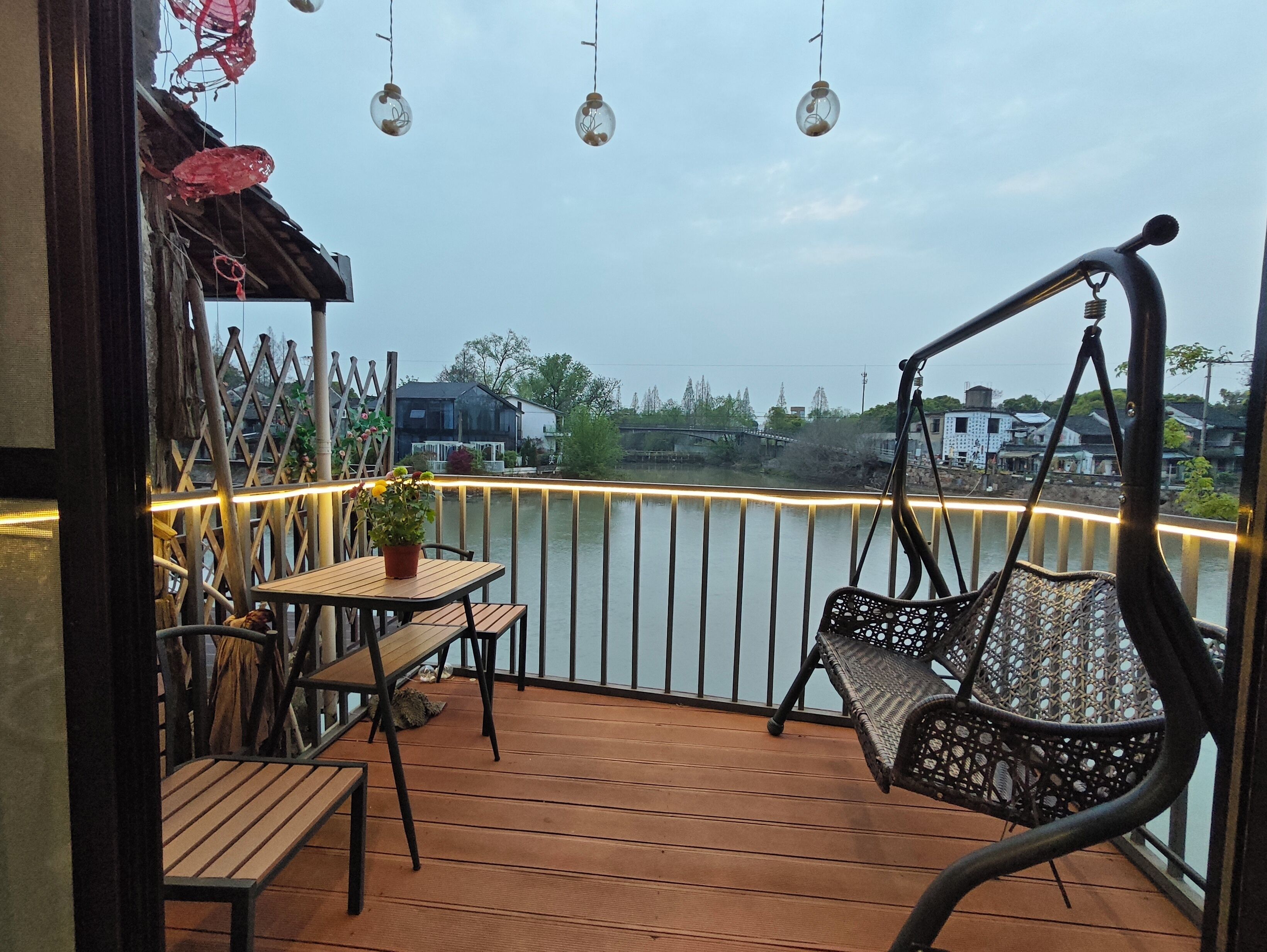 A wooden deck with a swing chair, a table with a potted plant, and a railing with lights.