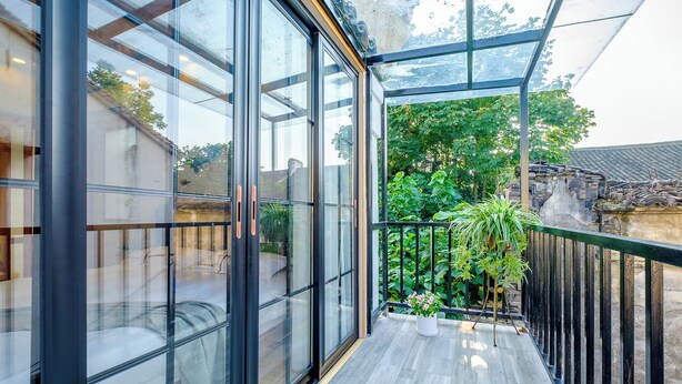 En moderne balkon med store glasdøre, trægulv og udsigt over en have samt traditionel arkitektur.