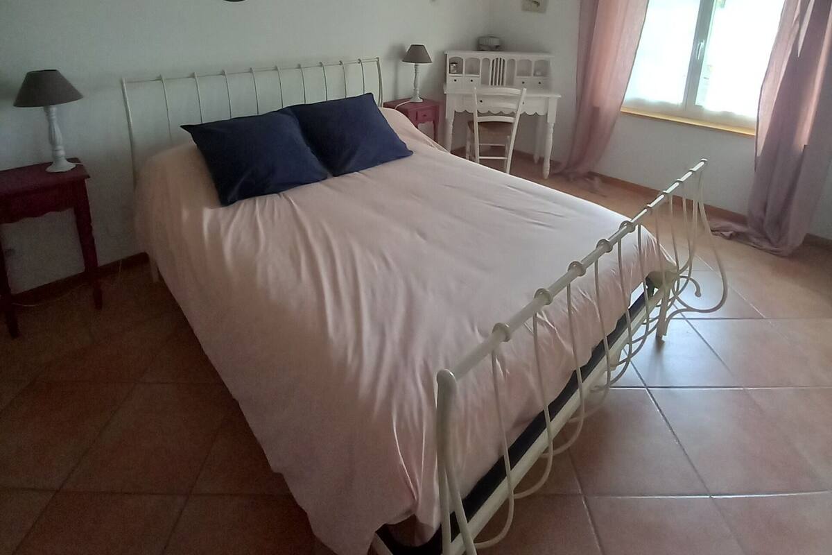 A bedroom with a bed, a bedside table, a lamp, a desk, a chair, and a window with curtains.