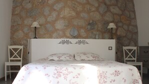 Un dormitorio con pared de piedra, cama con ropa de cama floral, dos mesitas de noche con lámparas y una silla de madera.
