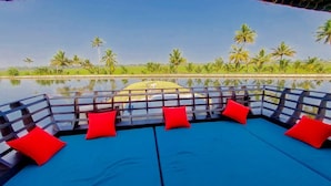 A poolside area with a blue cushioned seating arrangement and red cushions, overlooking a calm water body and palm trees. - Beachparadise  Daycruise houseboat (Ambalapuzha)