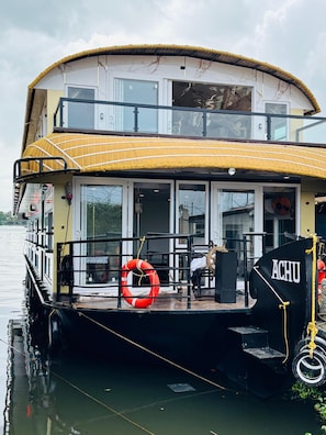 A houseboat with a two-level structure, featuring a deck area with a life ring, a steering wheel, and a sign labeled 'ACHU.'