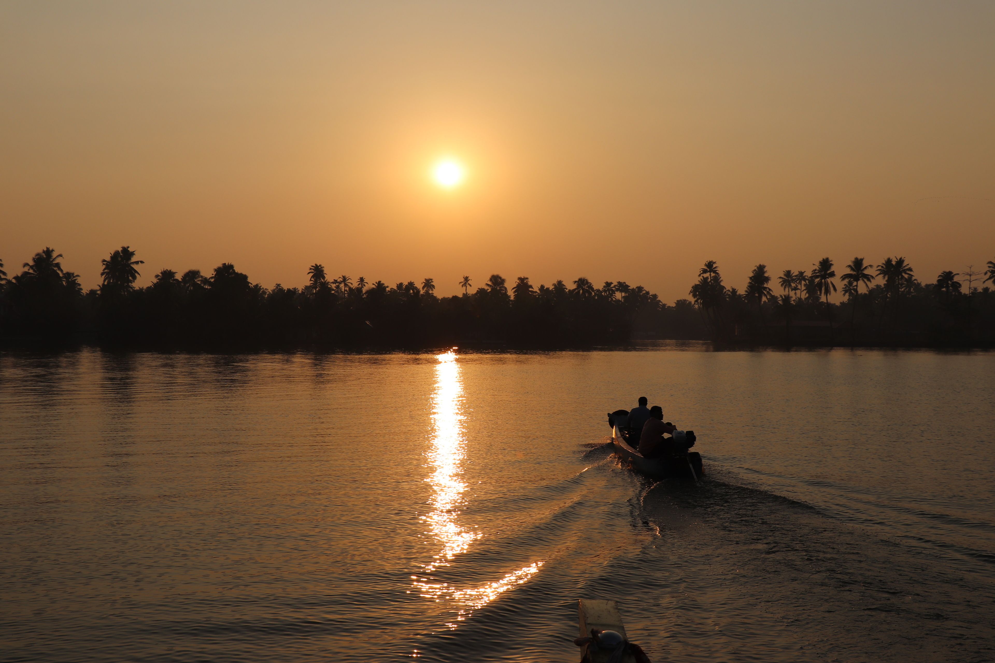 Atardecer sobre una superficie de agua tranquila con siluetas de palmeras y un bote con gente.