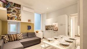 A modern living room with a grey sofa, a dining table set for four, and a kitchen area with white cabinets and a tiled backsplash.