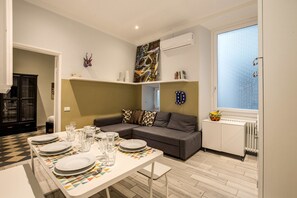 A modern living room with a dining area, a gray sofa, a patterned tablecloth, and a window with a view.