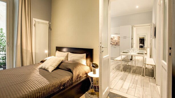 A bedroom with a bed, bedside table, and a view of the adjacent dining area.