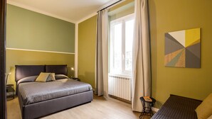Een slaapkamer met een groot bed, een kleiner bed, een raam met gordijnen, een radiator en een aan de muur bevestigd kunstwerk.
