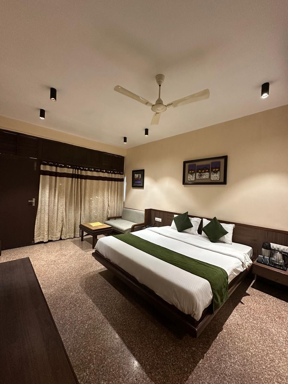 A hotel room with a large bed, a desk, a chair, a ceiling fan, and a nightstand with a coffee maker.